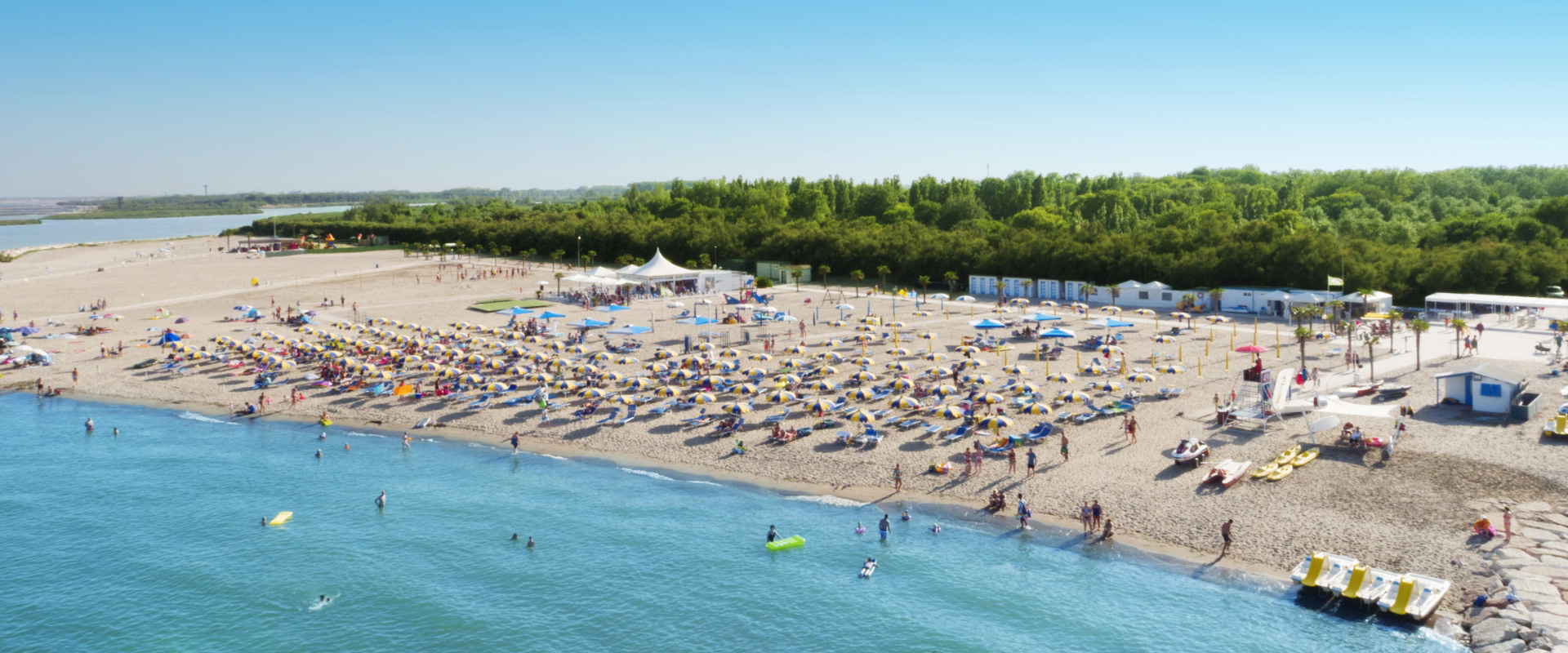 Spiaggia affollata con ombrelloni colorati e mare calmo, circondata da vegetazione verde.