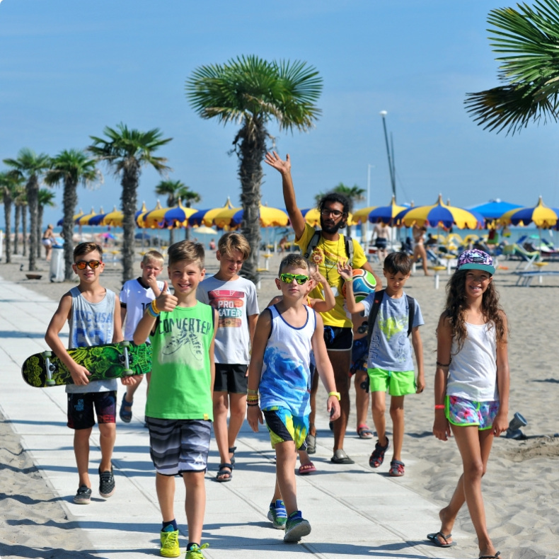 Gruppo di bambini felici cammina sulla spiaggia con palme e ombrelloni colorati.