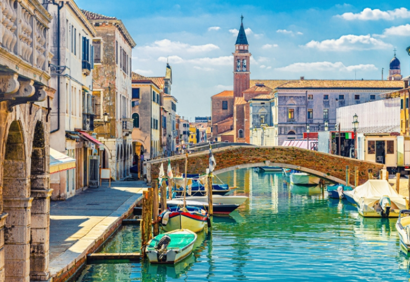 Canale veneziano con barche, ponte in mattoni e campanile sullo sfondo.