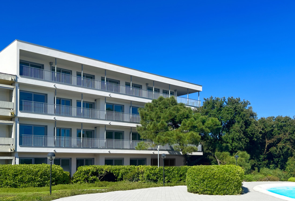 Edificio moderno con balconi, circondato da alberi e piscina, cielo limpido.