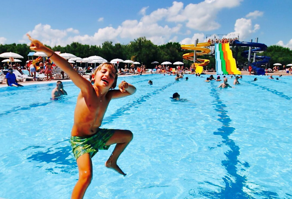 Bambino felice salta in piscina con scivoli colorati sullo sfondo.