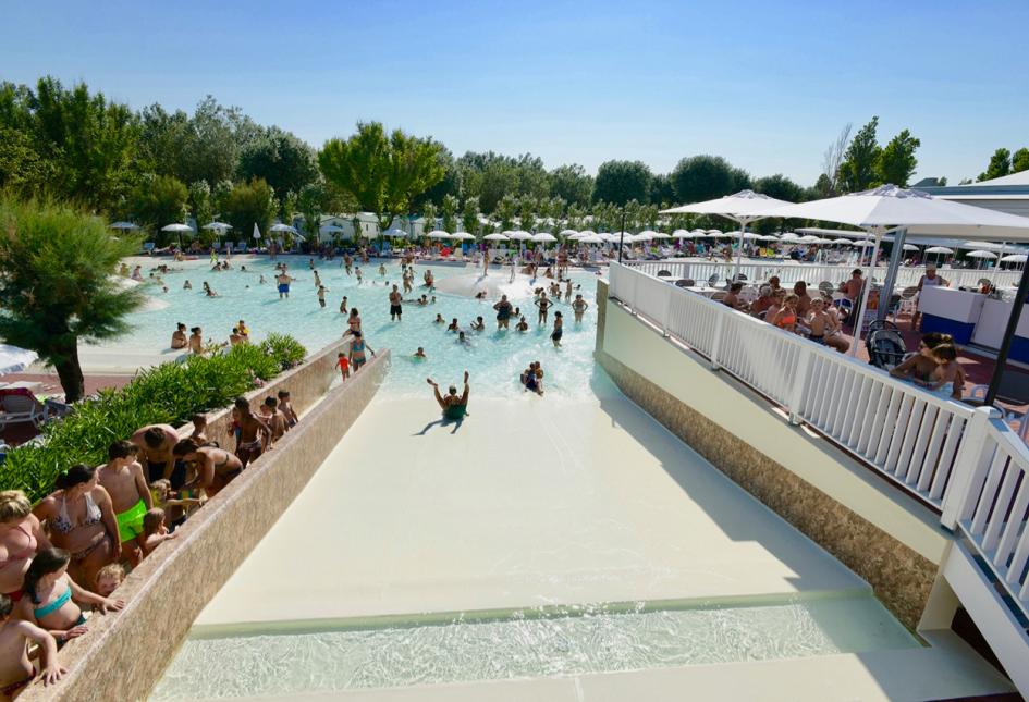 Piscina affollata con scivolo, ombrelloni e persone che si divertono al sole.