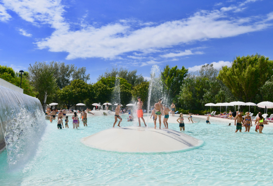 Piscina all'aperto con giochi d'acqua e persone che si divertono sotto il sole.