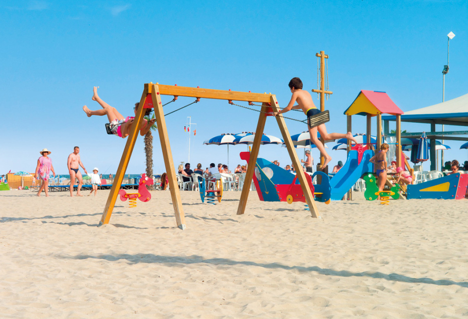 Spiaggia con parco giochi, bambini che giocano, adulti rilassati sotto ombrelloni.