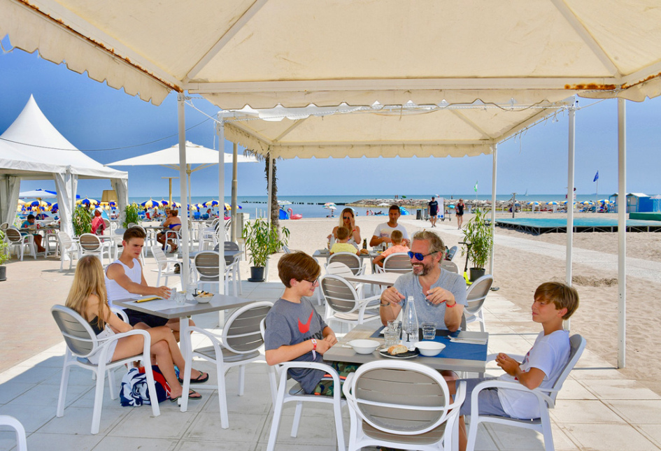 Famiglia pranza sotto il gazebo sulla spiaggia, con mare e ombrelloni sullo sfondo.