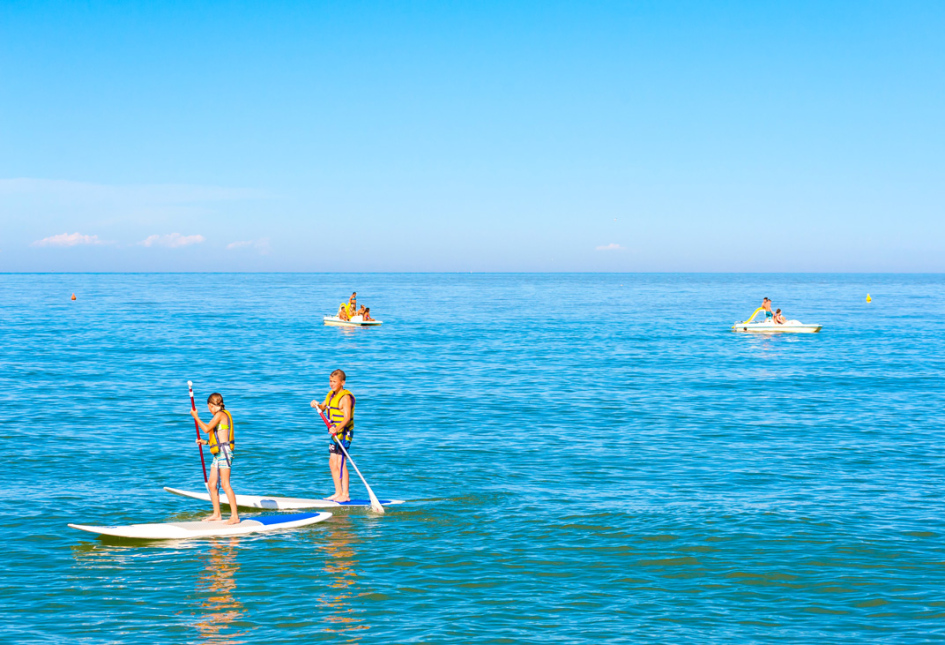 Persone fanno paddleboarding e pedalò in un mare calmo e soleggiato.